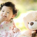 baby sitting on green grass beside bear plush toy at daytime